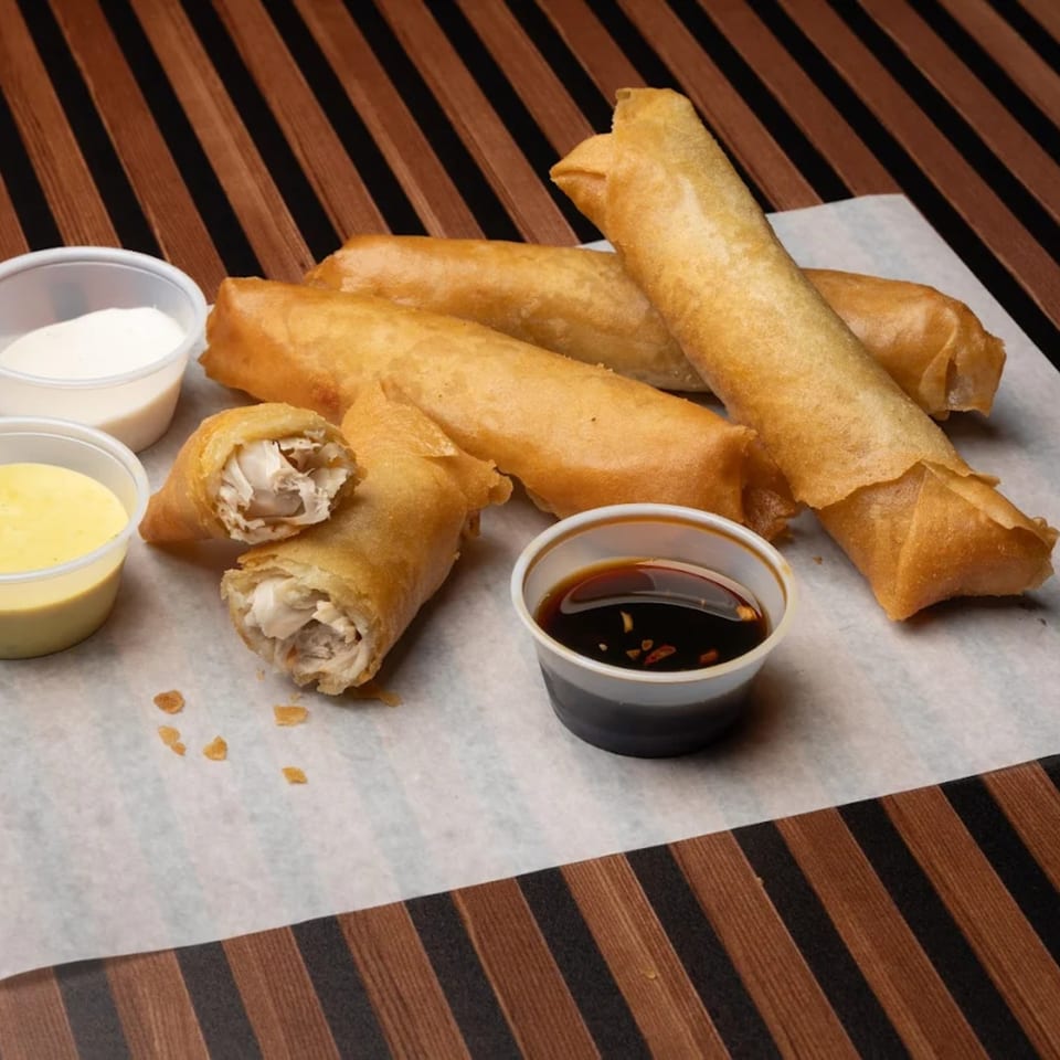 Crispy fried spring rolls with three dipping sauces on a white paper-lined wooden surface.
