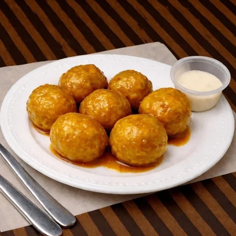 Seven golden-brown meatballs covered in sauce on a white plate with a side of dipping sauce.