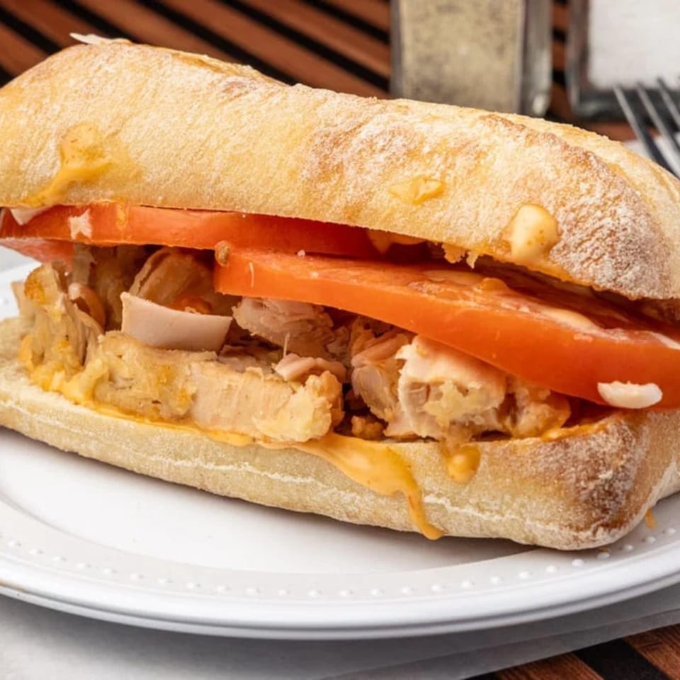 Close-up of a chicken and tomato sandwich on ciabatta bread with melted cheese, on a white plate.